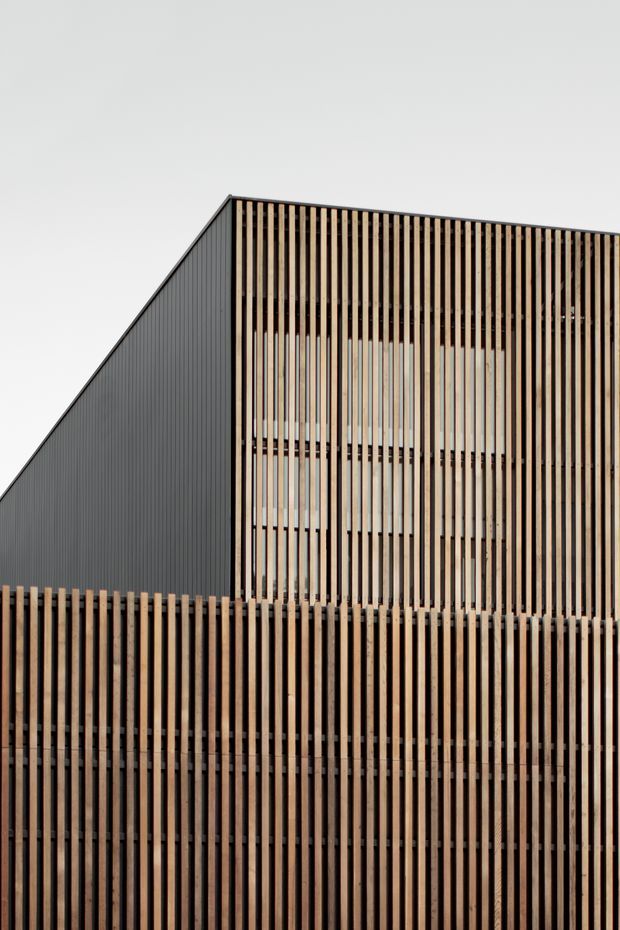 Close-up exterior view of a contemporary building with a strongly geometric, asymmetrical roofline. The facade combines dark vertically ribbed cladding with extensive vertical timber battens screening windows and upper-level glazing. A matching timber privacy or boundary screen dominates the foreground, creating a layered, rhythmic appearance.