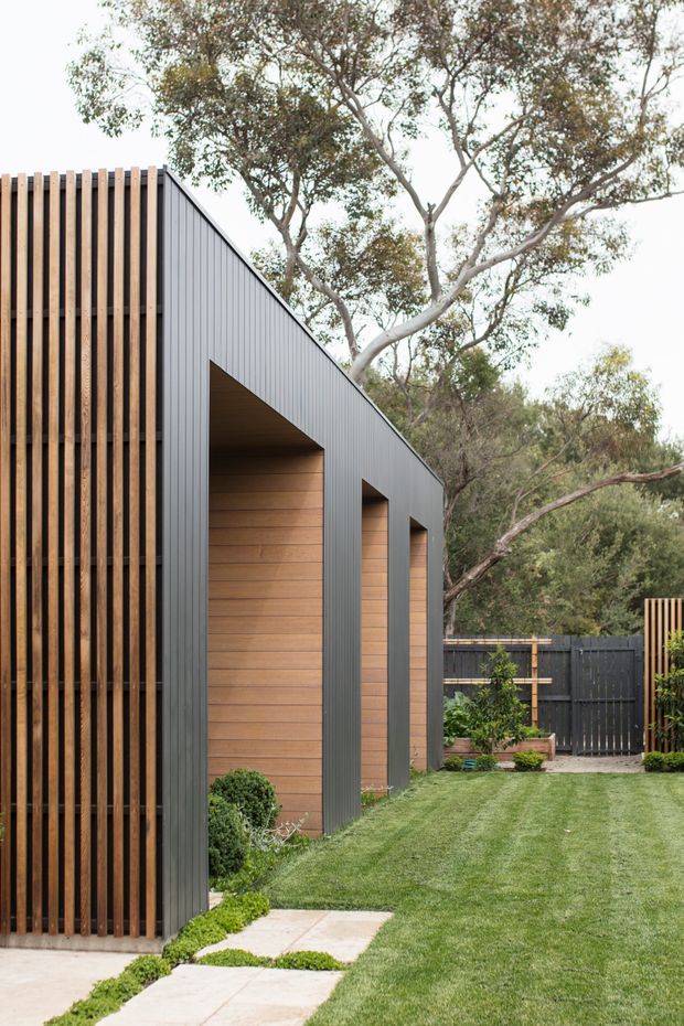 A contemporary landscaped residential exterior featuring a long, sharply pitched dark grey metal-clad structure with repeated recessed openings lined in warm horizontal timber. The foreground has a manicured lawn, pale concrete stepping path, clipped shrubs, timber screening and dark fencing, set among mature trees.