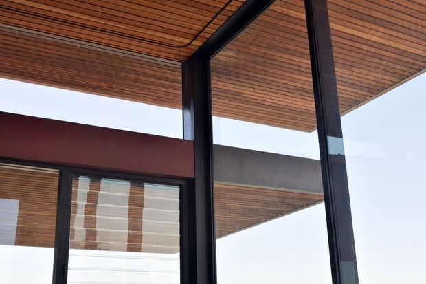 Close-up architectural view of a modern building corner with extensive floor-to-ceiling glazing framed in dark metal. Warm timber slat soffits and ceiling panels extend across the covered exterior, with strong horizontal and vertical lines, a deep burgundy wall band, and reflected timber surfaces visible in the glass.