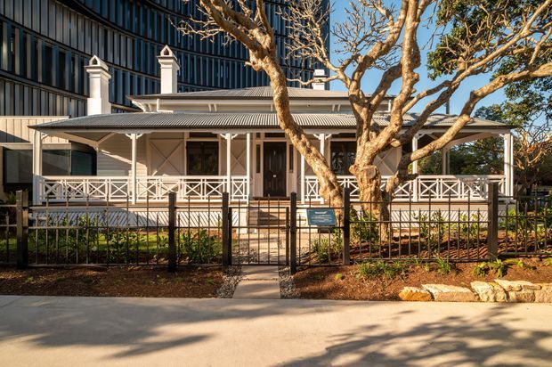 Street-facing view of a restored traditional timber veranda building with cream-painted columns, decorative cross-braced balustrades, corrugated metal roof, twin chimney forms, and a dark contemporary multi-storey building behind it. A mature leafless tree, landscaped front garden, black metal fence, and paved footpath frame the facade.