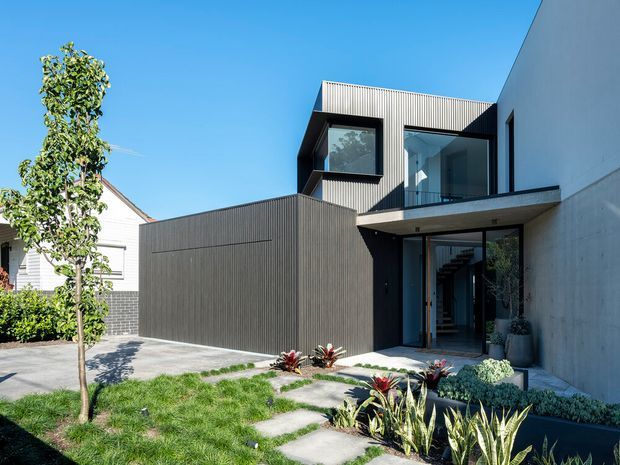 Contemporary multi-level residential facade with dark vertical cladding, smooth pale concrete walls, large glazed openings, a projecting upper-floor balcony, and a sheltered entry. A landscaped front garden with stepping-stone paving and low planting leads toward the entrance.