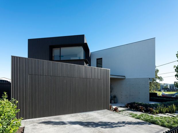Street-facing view of a contemporary multi-level residence with a stark geometric form. The building combines dark vertical slatted screening and cladding with smooth white upper walls and a raw concrete lower section. A broad concrete driveway and landscaped garden sit in front, with a partially concealed upper-level window and balcony-like opening behind the screen.