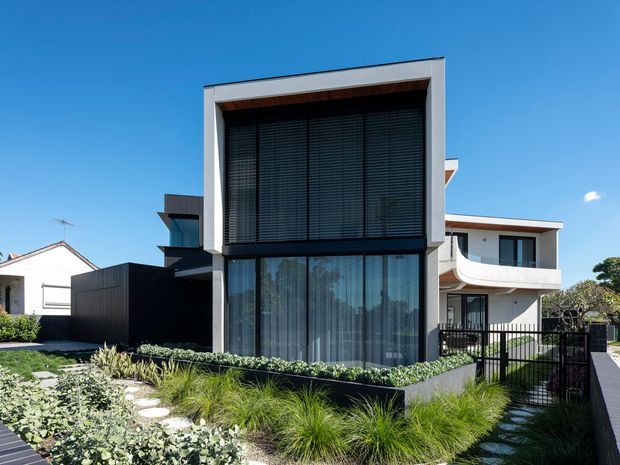 Street-facing view of a contemporary multi-storey residence with a strong rectilinear facade, large glazed openings, dark horizontal blinds, white rendered framing, and dark metal cladding. The front garden uses layered ornamental grasses and low planting, with a black metal gate and fencing. A neighbouring dwelling is visible beside the property under a clear blue sky.