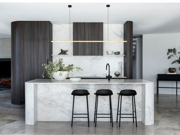 Contemporary kitchen featuring a large white marble-look island with integrated sink and black tapware, three black bar stools, dark vertical-grain timber cabinetry, open shelving, and a slim linear pendant light. The space has polished concrete flooring, white walls, and minimalist styling.