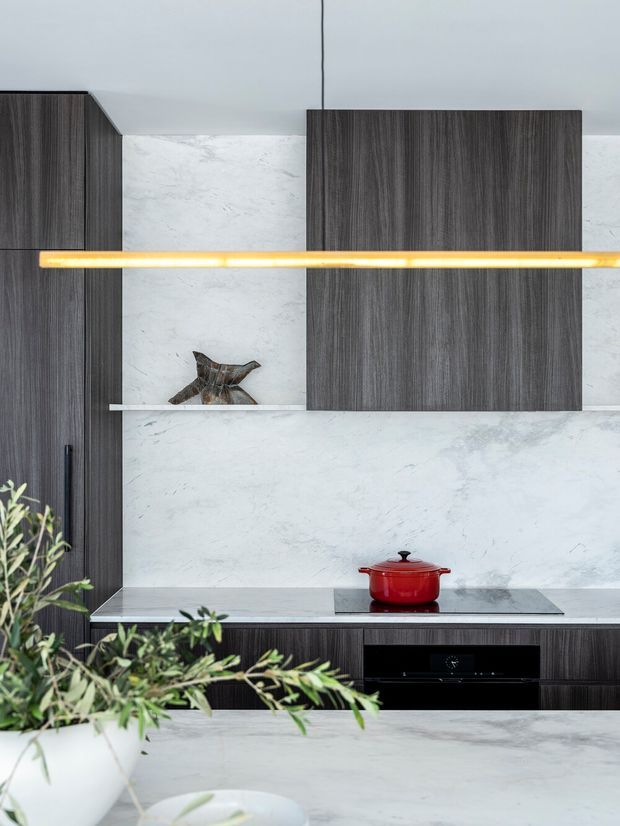 Contemporary kitchen interior featuring dark vertically grained timber cabinetry, a pale veined stone backsplash and benchtop, integrated appliances, a long warm linear pendant light, and a red Dutch oven on the cooktop. A large pale stone island occupies the foreground, with a leafy plant adding a natural accent.