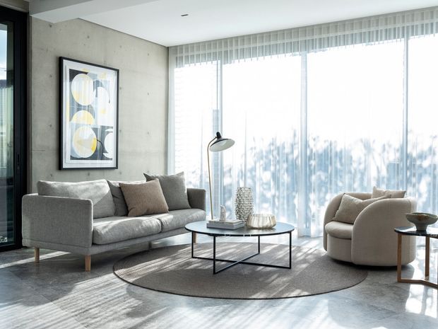 Bright contemporary living room with a grey sofa, sculptural cream lounge chair, round black-framed coffee table, large neutral rug, abstract wall art, concrete-look feature wall, and expansive floor-to-ceiling glazing with sheer blinds. The space has abundant natural light and a restrained neutral palette.