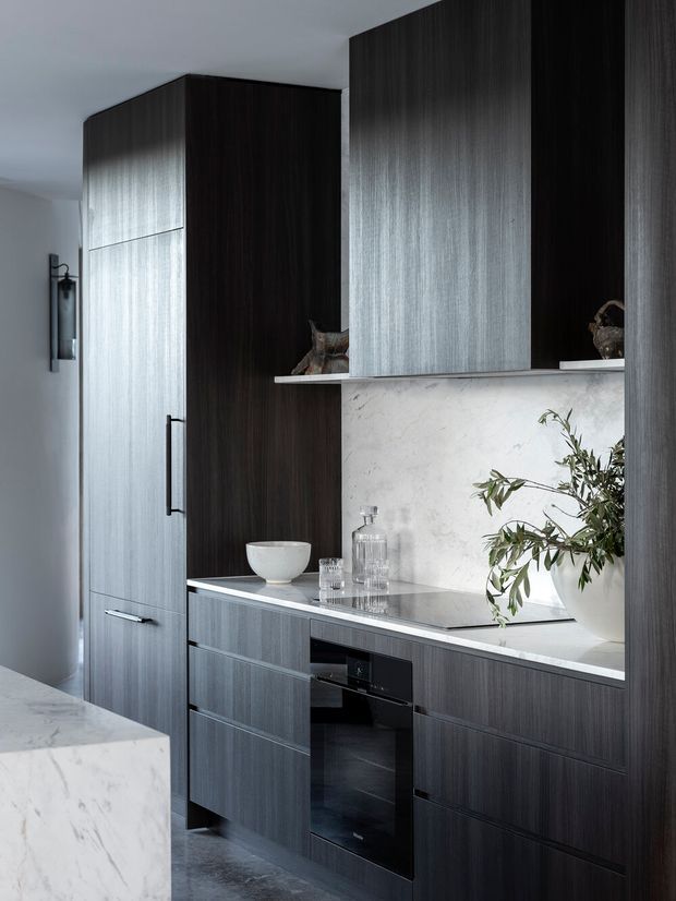 A refined contemporary kitchen with tall dark timber-grain cabinetry, integrated appliances, pale stone benchtops and splashback, open shelving, and minimal black hardware. A marble-look island edge is visible in the foreground, with restrained decorative objects and greenery adding softness.