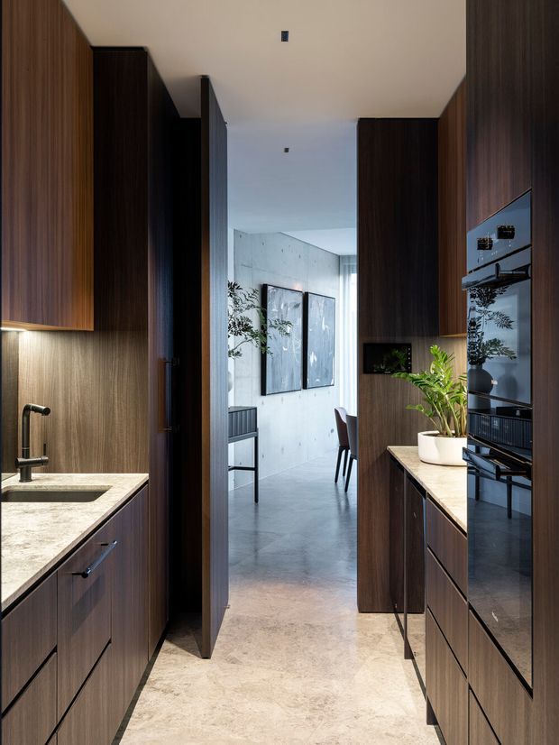 A narrow contemporary kitchen corridor with dark timber cabinetry on both sides, pale stone benchtops, integrated black appliances, and a stainless or dark metal tap. The kitchen opens toward a bright dining or living area with concrete-look walls, polished grey flooring, artwork, and a small table. Warm under-cabinet lighting and a potted plant add contrast to the restrained palette.