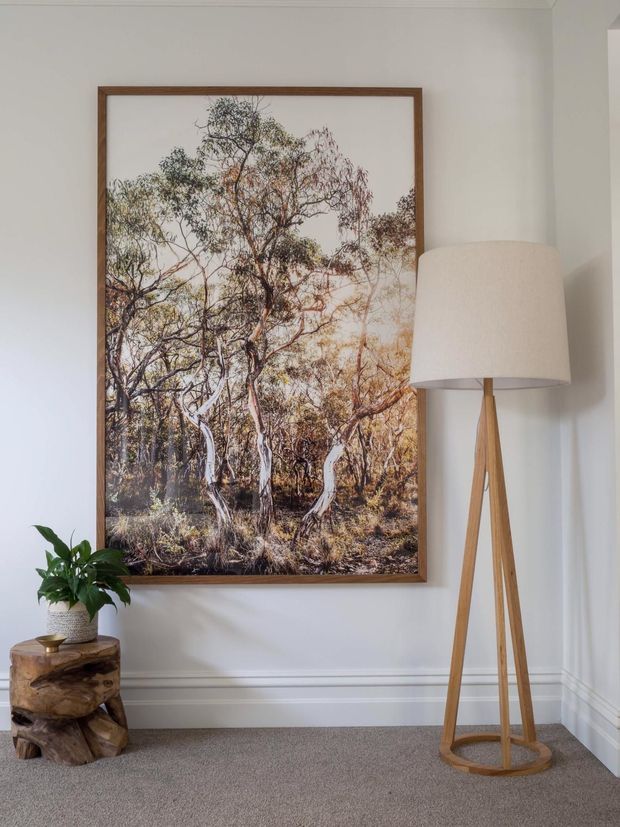 A calm, refined interior corner with a large framed Australian bush landscape artwork on a white panelled wall. A tall wooden tripod floor lamp with a cream fabric shade stands beside it, while a potted leafy plant sits on a sculptural natural timber stump table. The room has grey carpet, white decorative wall moulding, and a restrained neutral palette with warm timber accents.