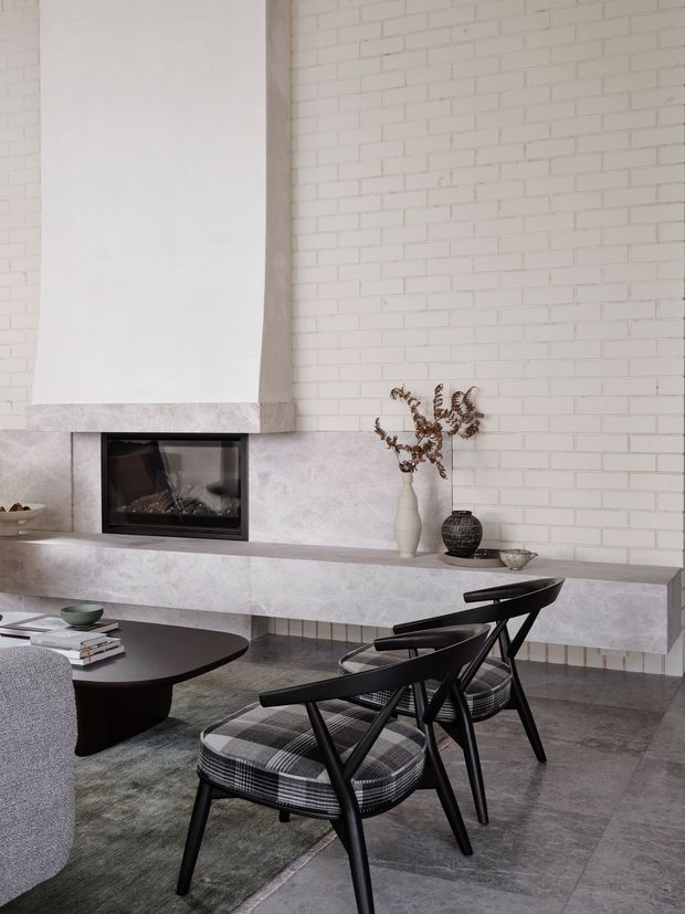 A refined contemporary living room centered on a long built-in fireplace wall. The room features pale painted brickwork, a large sculptural fireplace surround and hearth in pale stone, dark grey stone-look flooring, a low black coffee table, and black timber lounge chairs with patterned upholstery. Neutral styling, dried botanical decor, and restrained furnishings create a sophisticated, calm interior.