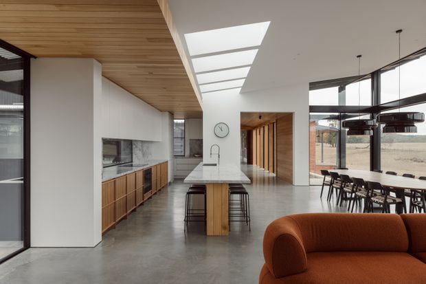 Open-plan contemporary interior featuring a kitchen, central island, dining area and living zone. The space has polished concrete flooring, extensive timber cabinetry, a timber-lined ceiling, white walls, skylights and large black-framed windows. A warm rust-orange sofa occupies the foreground, while dark pendant lights hang over the dining table.