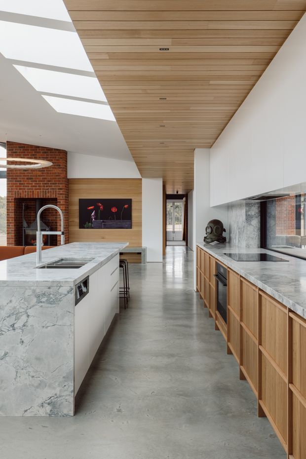 A bright, contemporary open-plan kitchen with a large marble-clad island, integrated sink and tap, timber cabinetry, polished concrete flooring, white overhead joinery, and a dramatic timber-lined ceiling. A red brick feature wall, black-framed fireplace, and skylight-like ceiling openings add contrast, while a dining or living area extends beyond the kitchen.