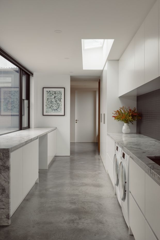 A bright, narrow contemporary utility kitchen and laundry corridor with white flat-panel cabinetry, grey stone benchtops, a dark tiled splashback, integrated laundry appliances, polished concrete flooring, large glazing, and a skylight. A central hallway and framed artwork are visible beyond the space.
