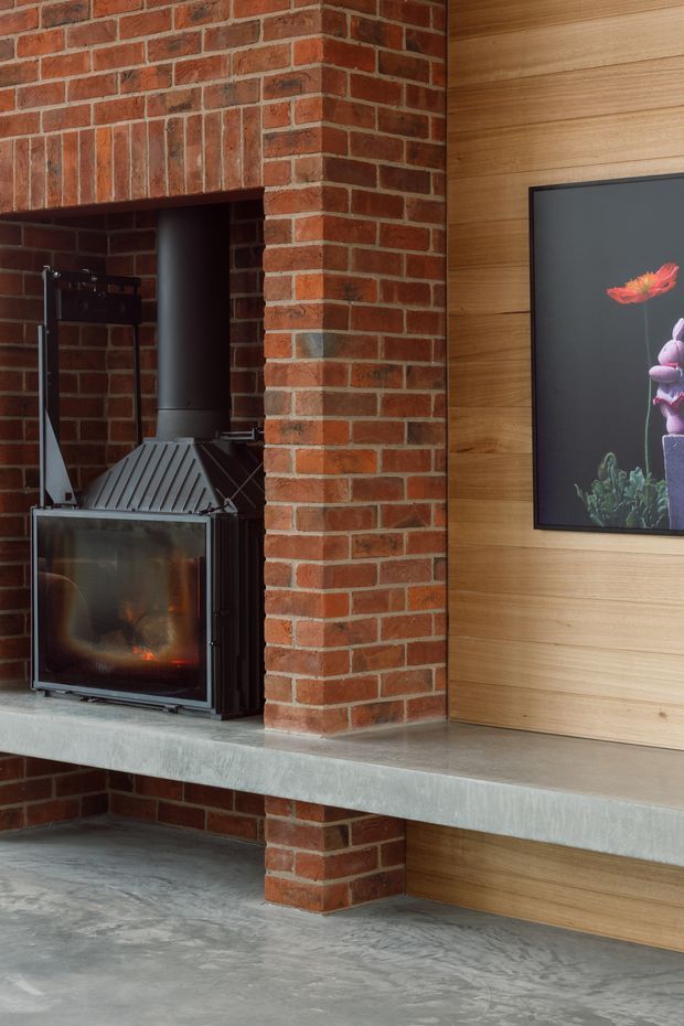 Close-up interior view of a modern fireplace built into a red brick wall, with a black wood-burning stove, concrete hearth shelf, and black flue. The adjacent wall is clad in horizontal warm timber boards and includes a framed floral artwork. A polished grey concrete floor continues below.