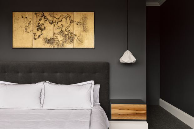 A moody, refined bedroom with a large upholstered charcoal-grey bedhead against a dark grey wall, crisp white bedding, a floating timber bedside table, and a faceted white pendant light. A framed sepia-toned artwork hangs above the bed, while dark carpet, deep wall tones, and white skirting create a high-contrast, intimate atmosphere.