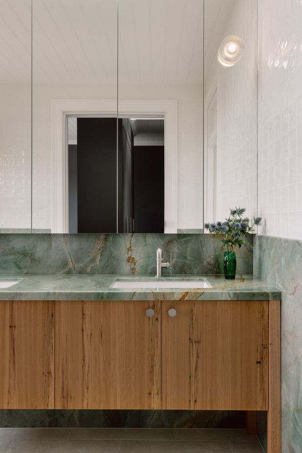 A refined bathroom vanity with a wall-mounted timber cabinet, integrated rectangular basin, and brushed metal tapware. Green veined stone forms the benchtop and backsplash, while glossy white tiled walls, a large mirror, recessed ceiling light, and a small green glass vase complete the space.