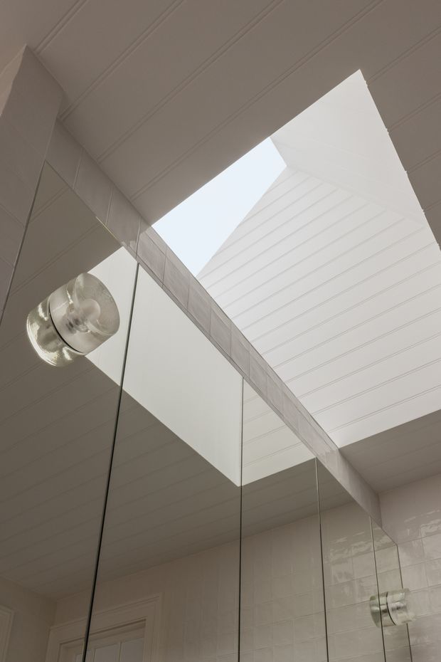 Upward-looking view of a bright, refined bathroom interior featuring a large skylight or roof opening, white painted tongue-and-groove ceiling boards, glossy white wall tiles, and tall mirrored surfaces. A clear glass pendant light with a visible reflection hangs near the ceiling, emphasizing the airy vertical volume and natural light.