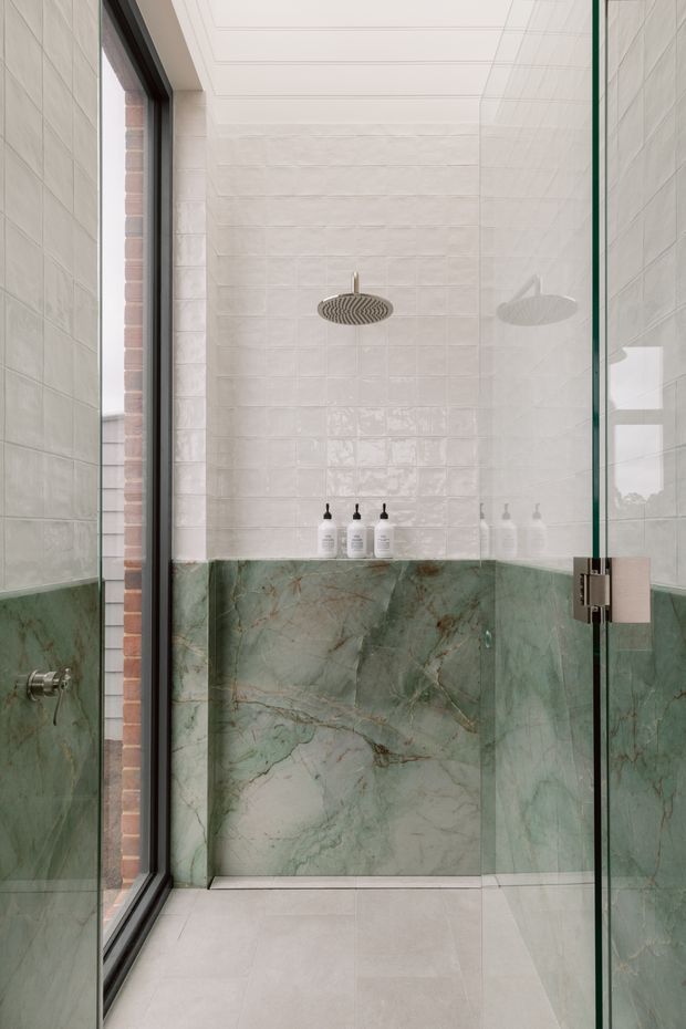 A refined walk-in shower enclosed by clear glass, featuring pale textured wall tiles, a green veined stone wainscot, a large round rainfall showerhead, dark-framed glazing, and light stone floor tiles. Minimal black-and-white toiletries sit on a narrow ledge, creating a clean, luxurious bathroom interior.