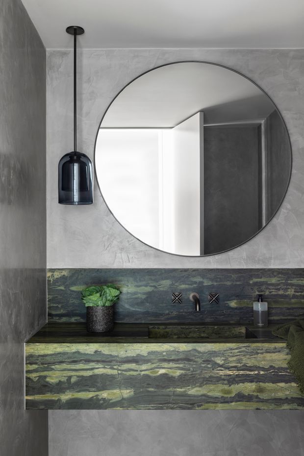 A compact, highly styled bathroom vanity area featuring a large round frameless mirror, dark pendant light, wall-mounted black tapware, and a dramatic green-and-charcoal veined stone backsplash and benchtop. The surrounding walls have a grey textured plaster finish, creating a moody, luxurious atmosphere. A small plant and soap dispenser sit on the vanity.