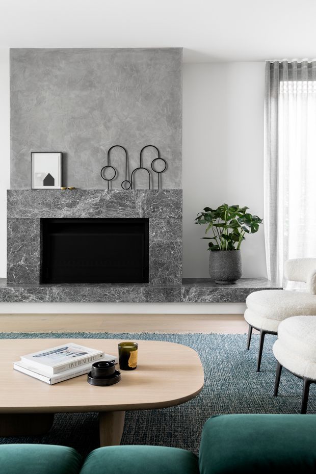 A refined contemporary living room centred on a dark veined stone fireplace with a textured grey feature wall and minimalist black sculptural decor. A low rounded light-wood coffee table, teal upholstered seating, boucle armchairs, blue-grey rug, sheer curtains, and a potted indoor plant create a calm, modern space with layered neutral and cool tones.