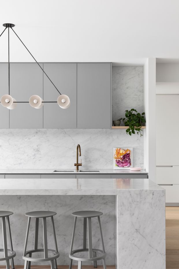 A refined contemporary kitchen with a large marble-clad island and integrated breakfast bar seating. The space features flat-front grey cabinetry, a matching marble splashback and benchtop, a brushed brass tap, and a minimalist linear pendant light. A small open shelf with greenery and artwork adds warmth, while pale timber flooring is visible beyond the kitchen.