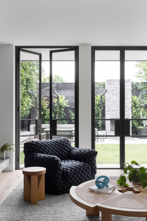 Bright contemporary living room with full-height black-framed glazing and open glass doors leading to a landscaped courtyard. A chunky dark boucle armchair sits beside a small timber side table, with a round timber coffee table, textured rug, and indoor plants in the foreground. The space has pale walls, abundant natural light, and a calm neutral palette with dark furniture accents.