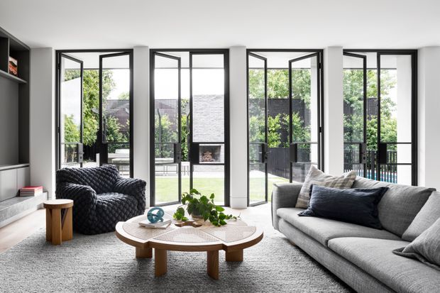 Bright contemporary living room with a large grey sectional sofa, textured dark armchair, round timber coffee table, integrated dark shelving and cabinetry, and full-height black-framed glazing opening to an outdoor terrace or courtyard. The room has white walls, a pale carpet, abundant natural light, and a restrained neutral palette with greenery and dark blue-grey accents.