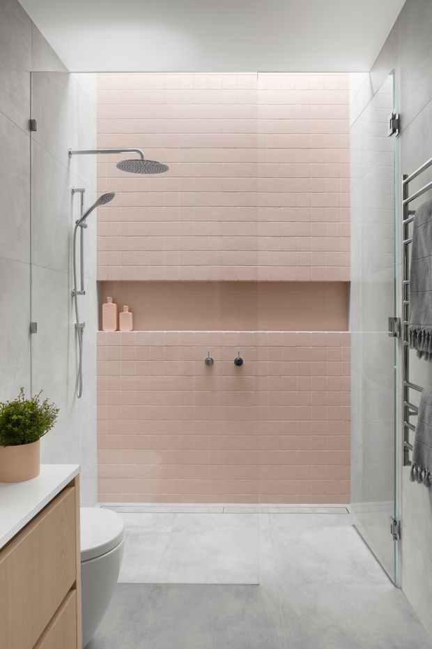 A contemporary bathroom featuring a walk-in shower enclosed by clear glass, with soft blush-pink small-format tiled walls and a recessed shelf. Large light-grey floor and wall tiles create a calm, cohesive backdrop. The shower has brushed metal fixtures, a handheld and overhead shower, and a wall-mounted towel warmer. A pale timber vanity with a white benchtop, toilet, and potted plant are partially visible in the foreground.