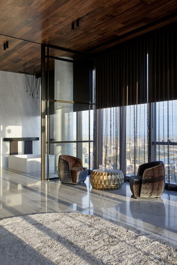 A luxurious high-rise living room with expansive floor-to-ceiling glazing and vertical dark blinds overlooking a cityscape. The room features a dark timber ceiling, polished light stone flooring, a pale textured rug, sculptural brown leather lounge chairs, and a metallic geometric coffee table. A minimalist fireplace and concrete wall are visible behind a glass partition.