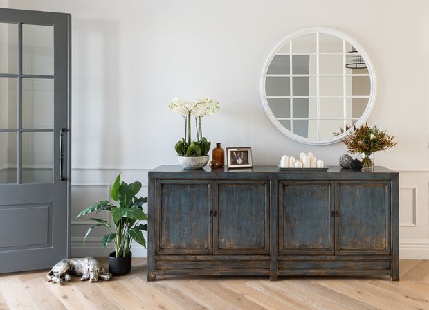 A refined interior vignette featuring a distressed dark wood sideboard with paneled doors, styled with plants, candles, framed artwork, and decorative objects. A large round white-paned mirror hangs above the sideboard, while a dark glazed door, white wall panelling, and light timber flooring complete the room. A small dog is visible on the floor.