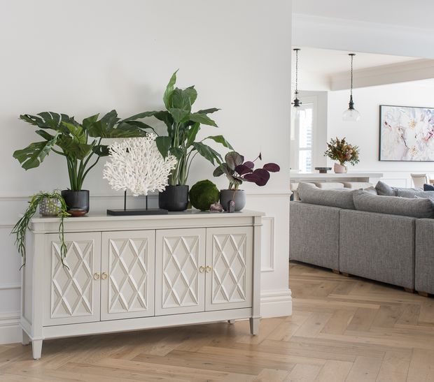 Bright, refined interior featuring a pale decorative sideboard with geometric diamond-panel doors, brass-toned knobs, and tapered legs. Large potted tropical plants and sculptural coral decor sit on top. The sideboard is positioned against white panelled walls, opening visually into a living room with a large grey sectional sofa, pendant lights, framed artwork, and light timber herringbone flooring.
