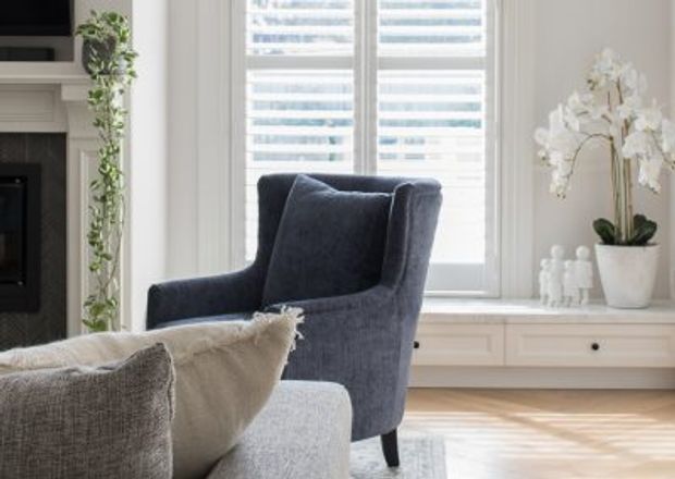 Bright, refined living room corner featuring a deep navy upholstered wingback armchair beside a light neutral sofa, a white fireplace surround, plantation shutters, built-in cabinetry, light timber flooring, and an orchid display. The room has a calm, classic-contemporary feel with abundant natural light.