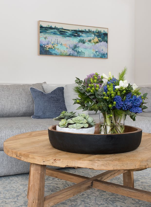 A styled living room vignette featuring a light grey upholstered sofa with a navy cushion, a chunky natural timber coffee table with crossed supports, and a large dark bowl holding succulents and a colourful blue-and-purple floral arrangement. A framed landscape artwork hangs on the pale wall behind the sofa, with a patterned rug beneath.