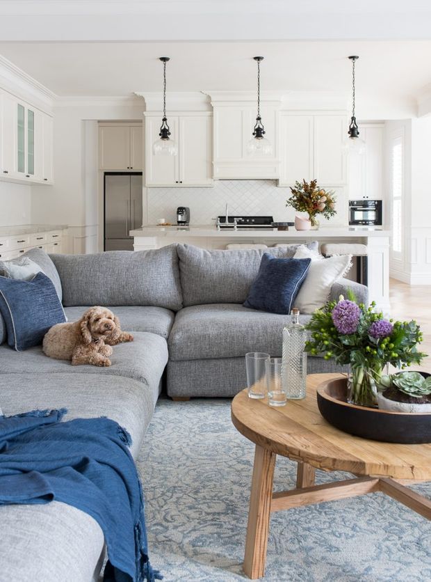 Bright open-plan living room with a large grey sectional sofa, blue cushions and throw, patterned pale rug, and a round timber coffee table with flowers and decorative objects. A white kitchen with shaker-style cabinetry, island bench, stainless-steel appliances, pendant lights, and tiled splashback is visible behind the seating area. The room has detailed cornices, abundant natural light, and a calm, refined coastal-inspired feel.