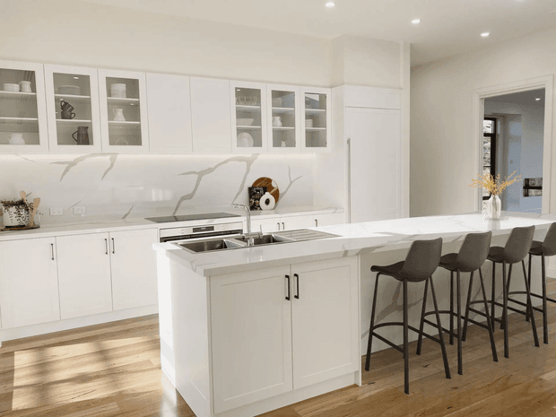 Bright, spacious contemporary white kitchen with a large central island incorporating a double sink and dishwasher, plus an extended breakfast bar with four dark upholstered stools. The room features white shaker-style cabinetry, glass-front display cabinets, integrated appliances, a white marble-look veined splashback and benchtop, recessed ceiling lighting, and warm timber flooring.