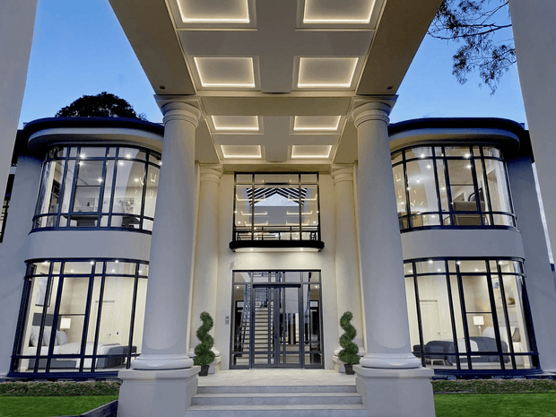 Formal symmetrical exterior entrance of a large multi-storey building, featuring a deep covered portico with illuminated recessed ceiling panels, monumental fluted classical columns, curved glass wings with dark window frames, a glazed central doorway, and an upper-level balcony. The facade combines classical architectural elements with contemporary extensive glazing, photographed at dusk.