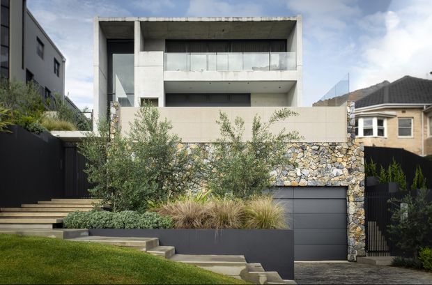Street-facing view of a contemporary multi-level residence with a strong rectilinear concrete facade, recessed glazing, a broad upper balcony, and a dark garage door. The front boundary combines a pale rendered wall with a prominent irregular natural-stone feature wall, dark metal fencing, and layered landscaped planters with grasses and shrubs. Stepped concrete paths and lawn lead up to the entry, while neighbouring homes are visible on either side.