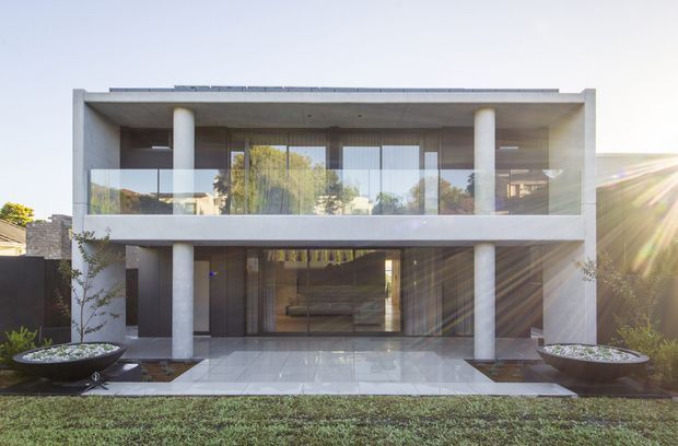 Front exterior view of a refined two-storey contemporary residence with a strong rectilinear form, flat roof, deep recessed upper-level balcony, extensive floor-to-ceiling glazing, and prominent cylindrical concrete columns. Pale stone or concrete paving leads to the glazed entry, with raised planter beds flanking the facade and a manicured lawn in front.