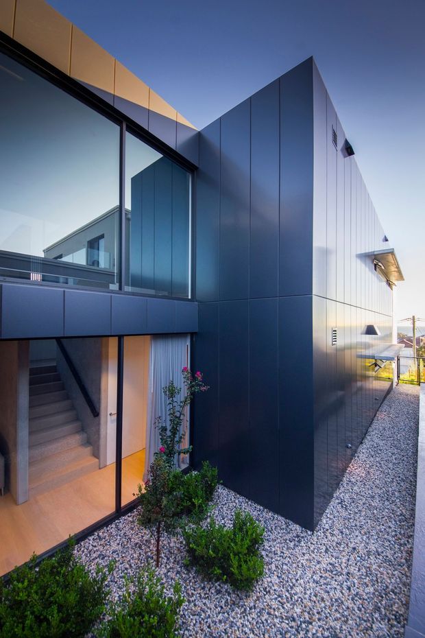 Exterior view of a contemporary multi-level residence with sharply articulated geometric volumes, dark blue-grey metal cladding, expansive black-framed glazing, and a recessed illuminated entry stair. A gravel-covered landscaped strip with low shrubs runs alongside the building, while balconies and projecting roof elements emphasize the angular form.