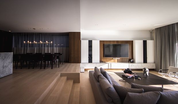 Open-plan contemporary living and dining area with a stepped timber floor, large sectional sofa, dark dining setting, and a built-in media wall with timber cabinetry and stone or light-toned surfaces. Full-height curtains and concealed ceiling lighting create a refined, warm, low-clutter interior.