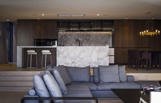 Open-plan contemporary living area with a large grey sectional sofa in the foreground, facing a dramatic marble kitchen island and splashback. Dark timber cabinetry, integrated appliances, a concealed bar or display area, dining table with dark upholstered chairs, layered split-level flooring, and a linear gold pendant light create a refined, moody interior.