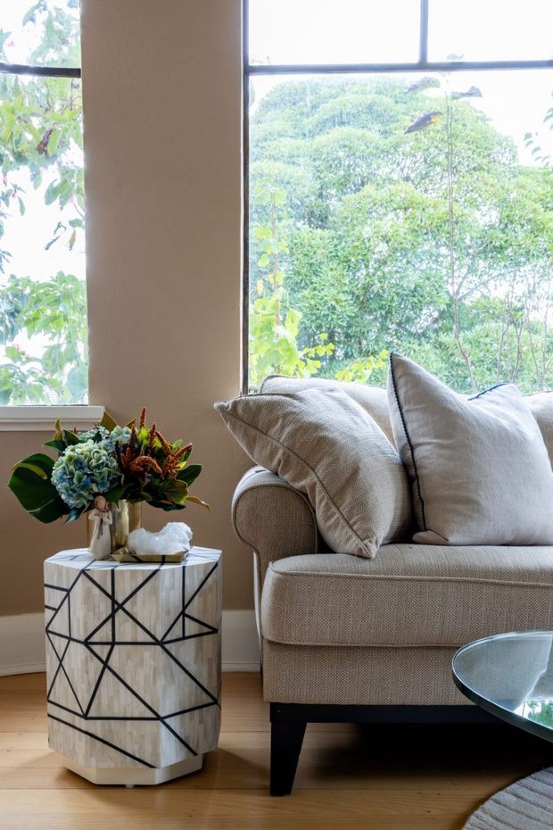 A close-up living room vignette with a textured beige sofa, layered neutral cushions, a geometric black-and-white side table with a floral arrangement, and a glass coffee table. Large black-framed windows provide abundant natural light and overlook leafy greenery. The room has timber flooring and a warm, refined neutral palette.