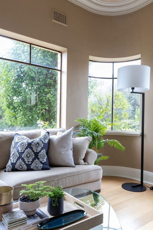 A bright, cozy living room corner with a light upholstered sofa, patterned navy-and-white cushion, potted plants, and a glass-topped coffee table. Large curved and rectangular steel-framed windows overlook dense greenery, while a tall black floor lamp stands beside the sofa. The room features warm beige textured walls, pale timber flooring, white decorative ceiling moulding, and a curved bay-window form.