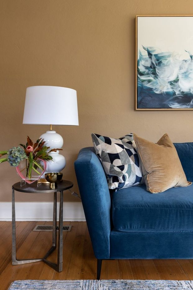 A warmly styled living room corner featuring a deep blue upholstered sofa with patterned black, white and blue cushions and a tan velvet cushion. Beside it is a slim black metal and glass side table with a white ceramic table lamp, floral arrangement and small decorative vessel. The room has warm taupe walls, timber flooring, white baseboards, a floor vent, and a framed abstract blue-and-white artwork. A patterned area rug is partially visible in the foreground.