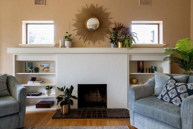A warm, traditionally styled living room centred on a white-painted fireplace with built-in open shelving on both sides. Two grey upholstered sofas frame the fireplace, with patterned cushions, timber flooring, decorative plants, a large sunburst mirror, and floral arrangements on the mantel. Two small windows and ceiling vents provide additional architectural detail.