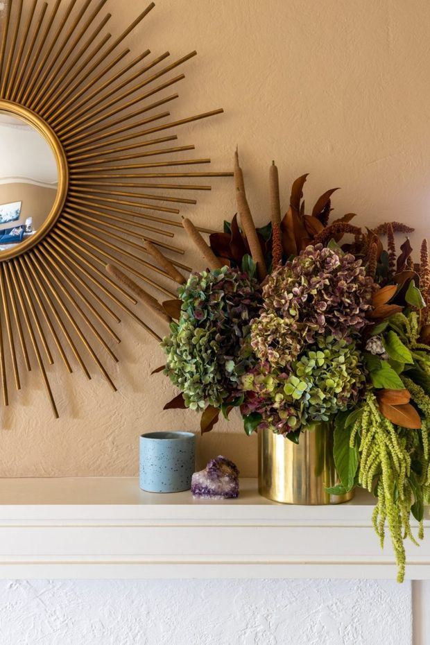 Close-up interior vignette of a decorative mantel or shelf against a warm textured beige wall. A large gold sunburst mirror dominates the left side, while a lush arrangement of dried and preserved flowers sits in a reflective brass-gold vase on the white moulded mantel. A small blue ceramic vessel and purple crystal accent complete the display. The mirror reflection reveals part of a nearby room, but the main subject is the styled mantel arrangement.