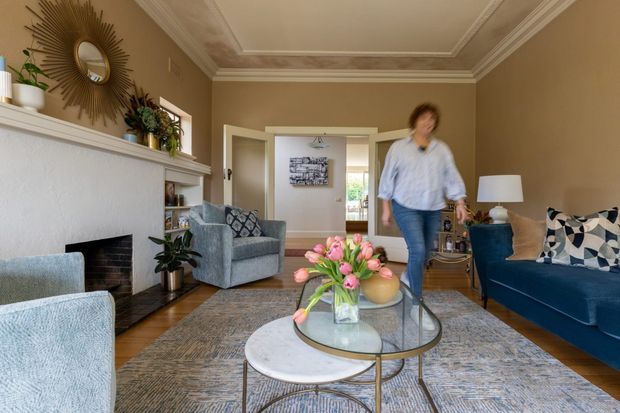 A warmly styled traditional living room with a white-painted fireplace and mantel, ornate ceiling cornices, neutral taupe walls, timber flooring, and layered area rugs. Blue upholstered armchairs and sofa surround nested round coffee tables, with patterned cushions, a glass vase of pink flowers, decorative mirror, plants, and table lamps adding colour and texture. A doorway leads to adjoining rooms in the background.