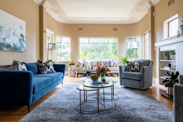A bright, traditionally detailed living room with warm beige walls, white decorative ceiling cornices and tray detailing, large black-framed windows, and a white fireplace surround with built-in shelving. The room is furnished with a deep blue sofa, grey armchairs, a pale sofa, nested glass coffee tables, a large textured grey rug, and patterned cushions. Abundant daylight and greenery visible through the windows create an inviting, refined atmosphere.