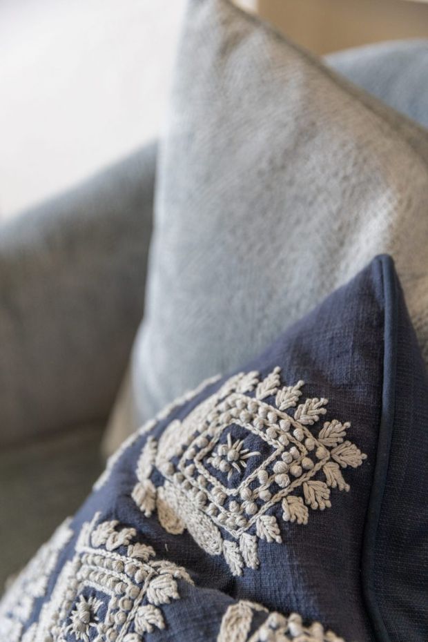Close-up detail of a navy-blue textured decorative cushion with intricate cream embroidered motifs resting against a light grey upholstered sofa. The image emphasizes soft textiles, layered neutral tones, and subtle tactile detailing in a softly lit interior.