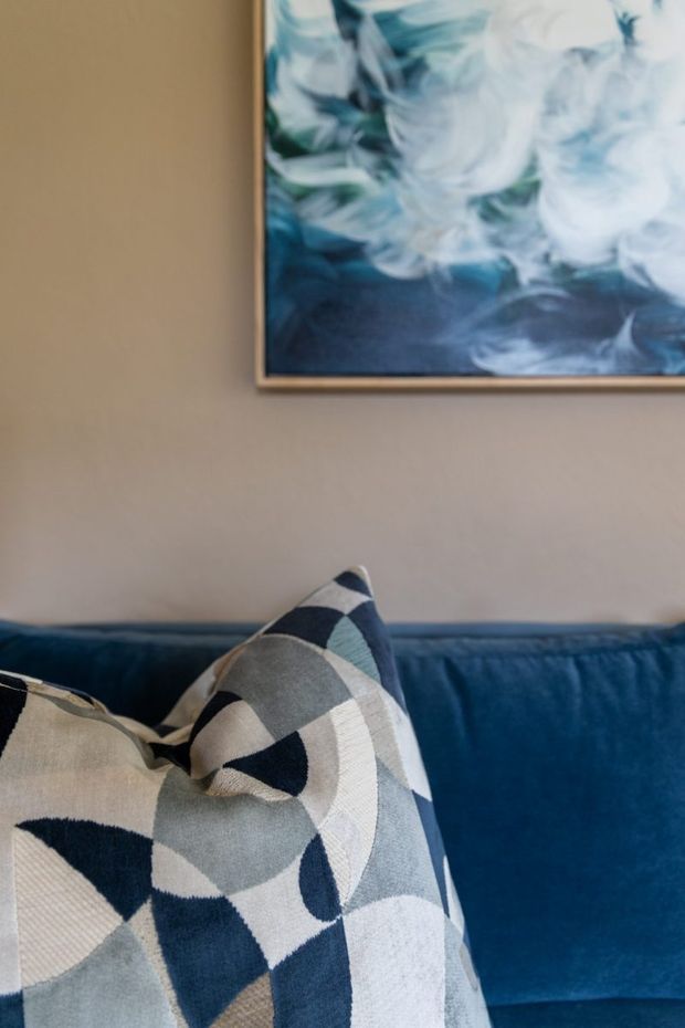 Close-up interior detail of a deep blue upholstered sofa with overlapping decorative cushions in a bold geometric pattern of navy, grey, cream and pale blue. A framed abstract artwork with swirling blue, white and green tones hangs on a warm taupe wall above the sofa.
